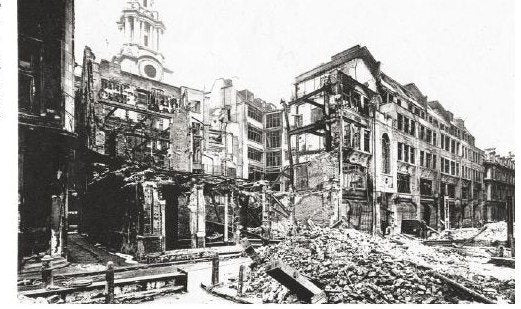 A black and white photograph of gutted roofless building shells and heaped rubble along a street, with a cathedral tower visible in the distance.